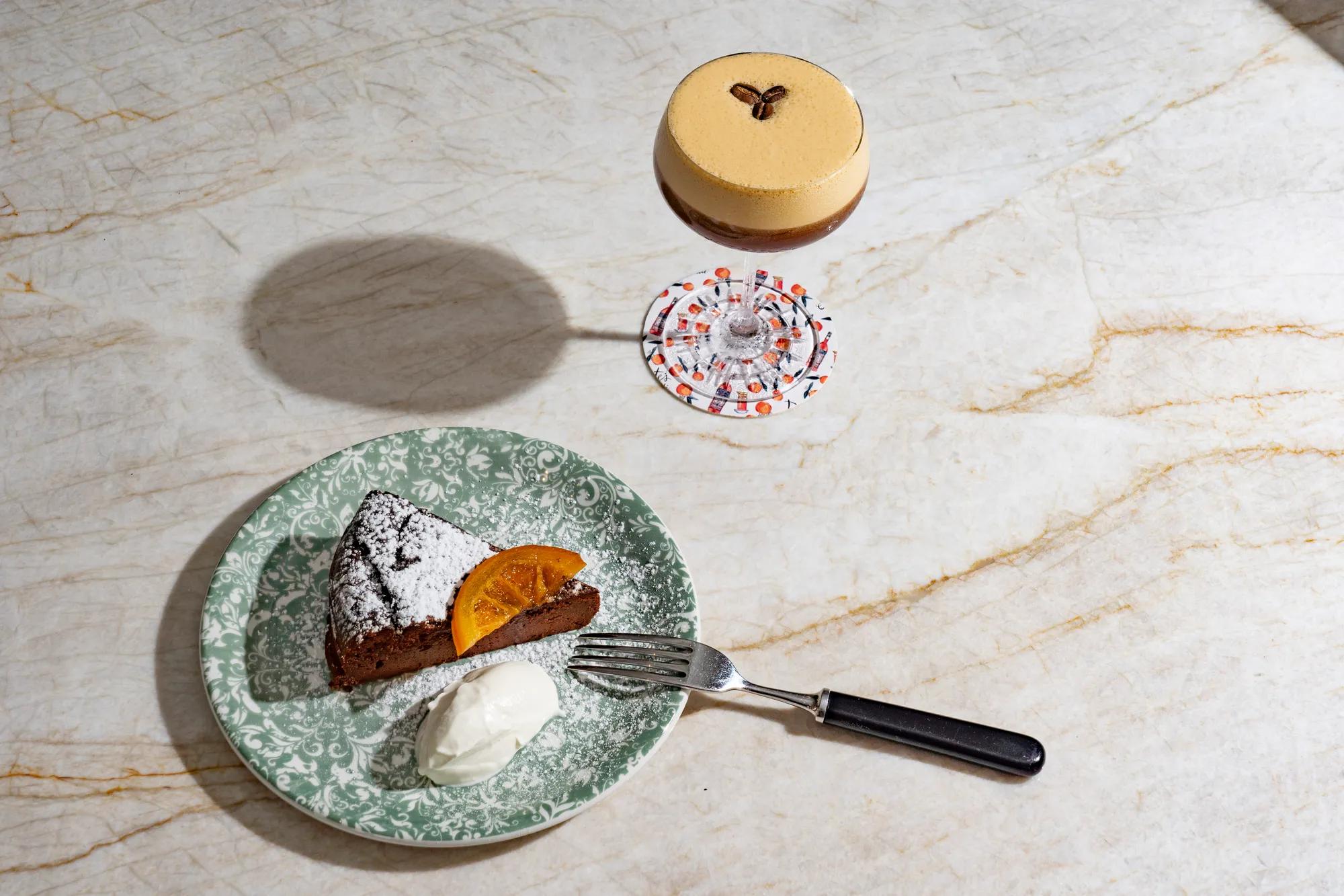 Slice of chocolate cake with cream beside an espresso martini cocktail on a marble surface.