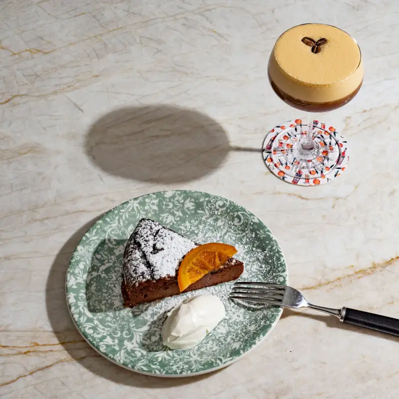 Slice of chocolate cake with cream beside an espresso martini cocktail on a marble surface.