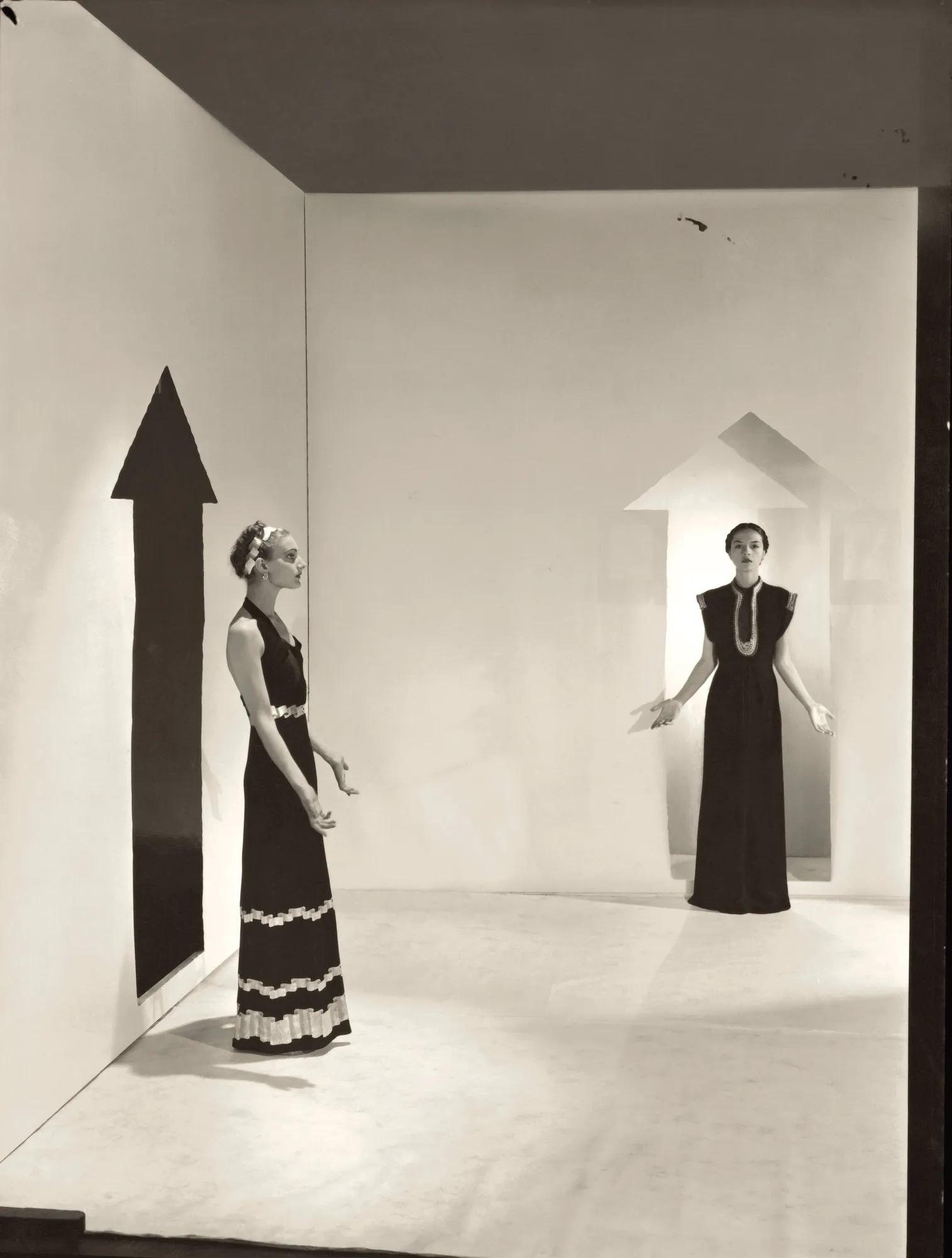 Two women in long dresses stand in minimalist room with painted arrows on walls, creating surreal scene.