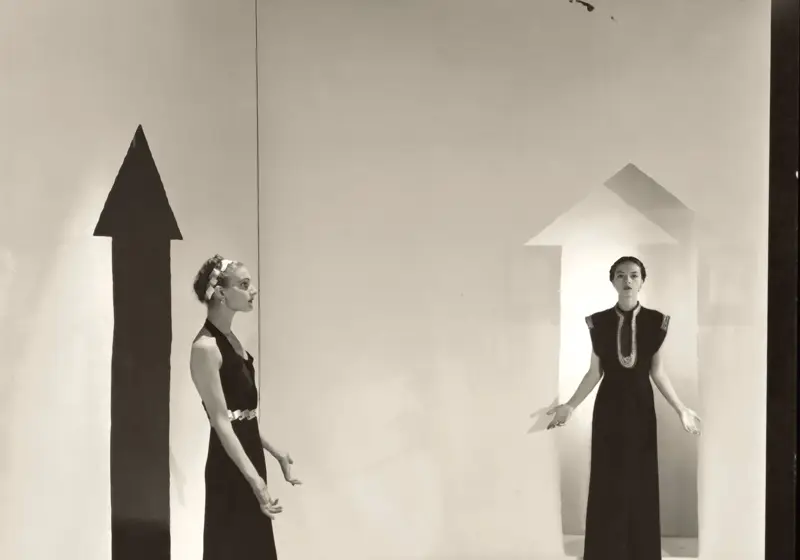 Two women in long dresses stand in minimalist room with painted arrows on walls, creating surreal scene.