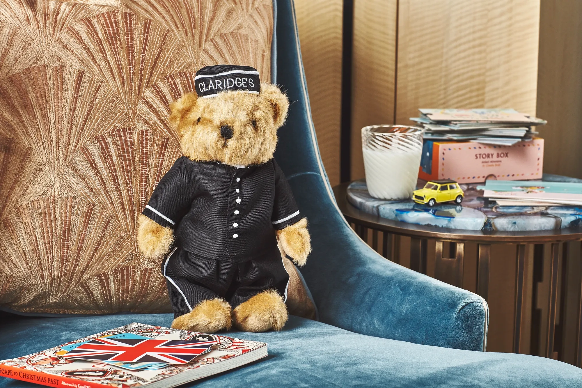 A Claridge’s teddy bear dressed in a black bellhop uniform sits on a blue velvet armchair beside a cushion, with a Union Jack book, toy car, and candle on a nearby table.