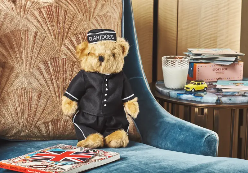 A Claridge’s teddy bear dressed in a black bellhop uniform sits on a blue velvet armchair beside a cushion, with a Union Jack book, toy car, and candle on a nearby table.