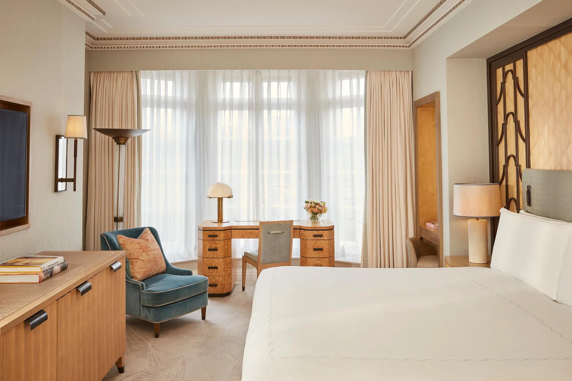 Elegant bedroom with a large bed, writing desk by a tall window, and classic décor featuring pale blue walls with gold trim and traditional furnishings.