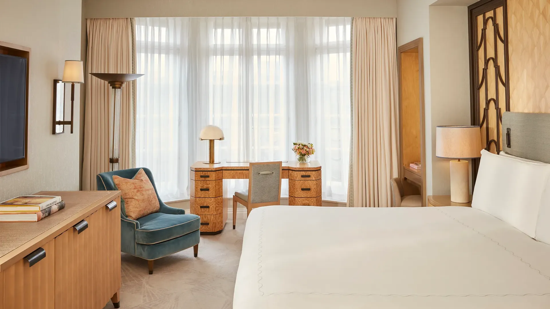 Elegant bedroom with a large bed, writing desk by a tall window, and classic décor featuring pale blue walls with gold trim and traditional furnishings.