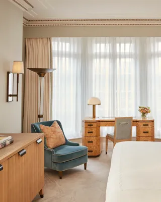 Elegant bedroom with a large bed, writing desk by a tall window, and classic décor featuring pale blue walls with gold trim and traditional furnishings.