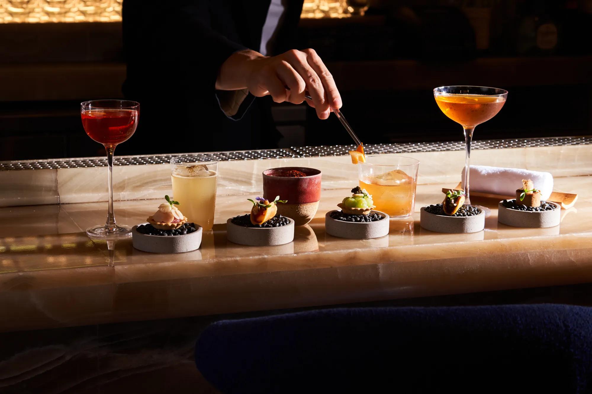 Bartender plating gourmet canapés with cocktails lined up on bar counter.