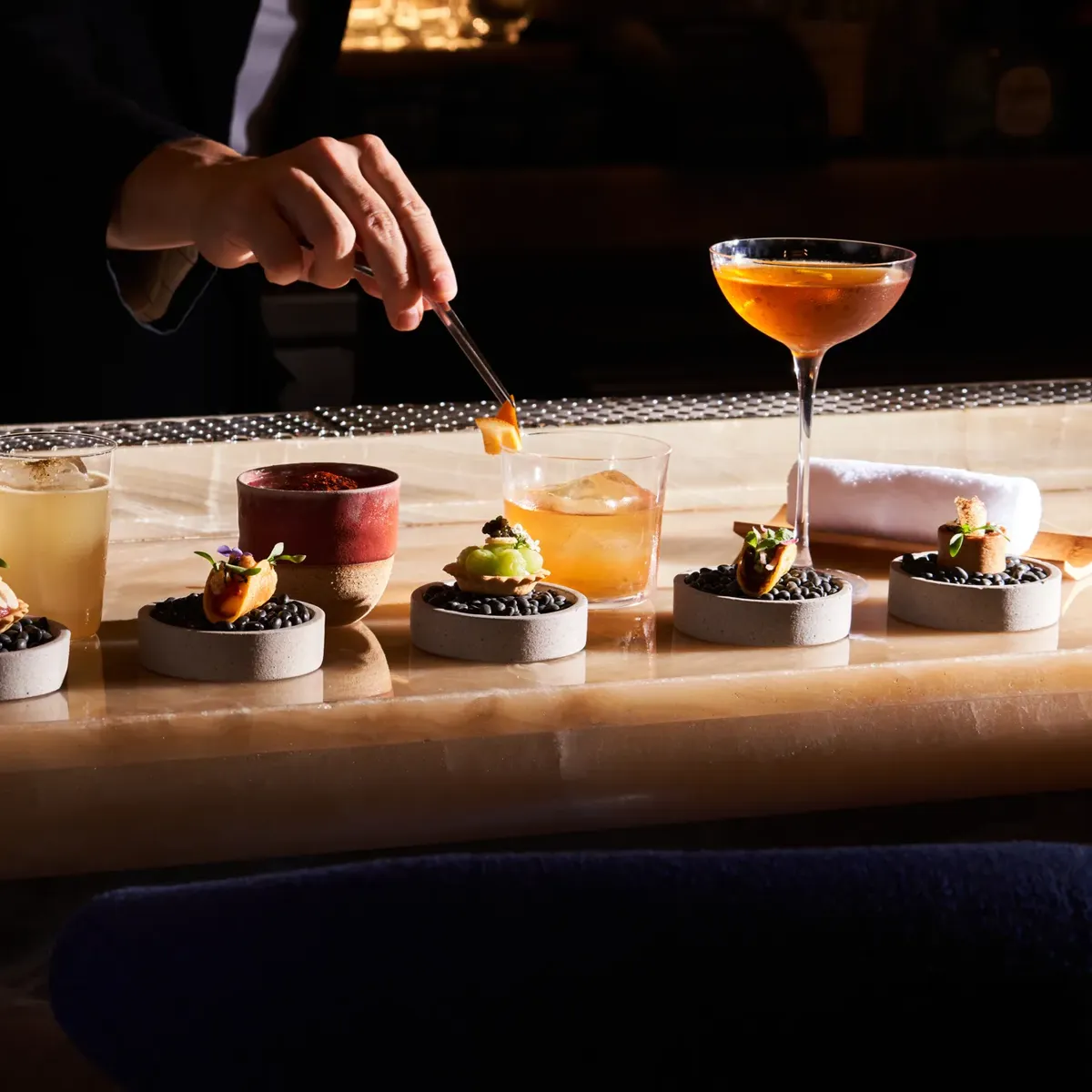 Bartender plating gourmet canapés with cocktails lined up on bar counter.
