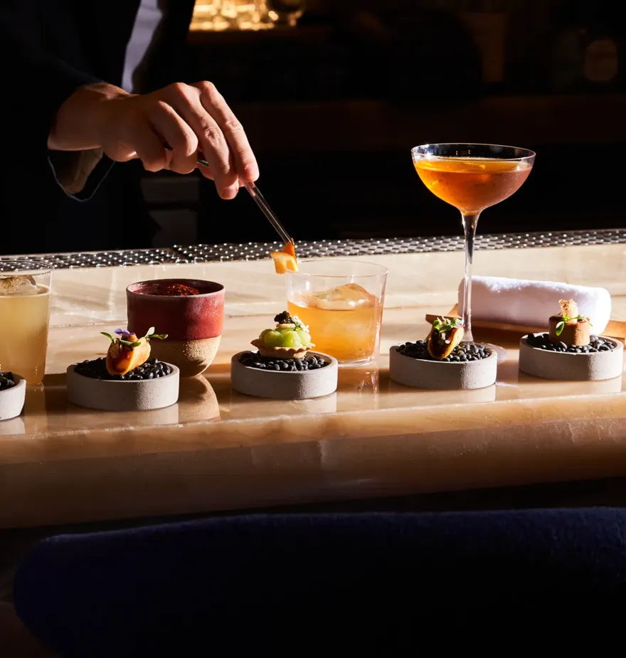 Bartender plating gourmet canapés with cocktails lined up on bar counter.