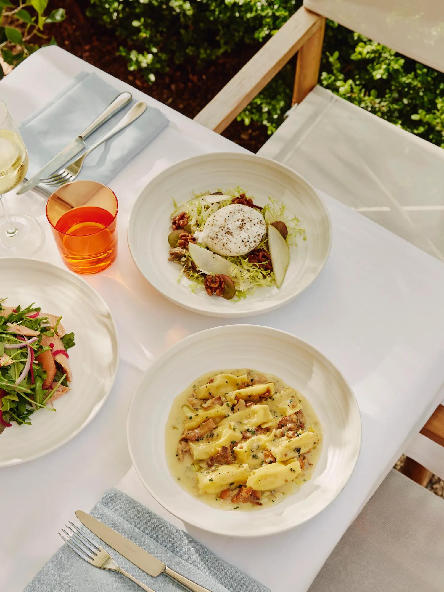Overhead of burrata and pear salad beside gnocchi in creamy sauce on a sunlit terrace table.