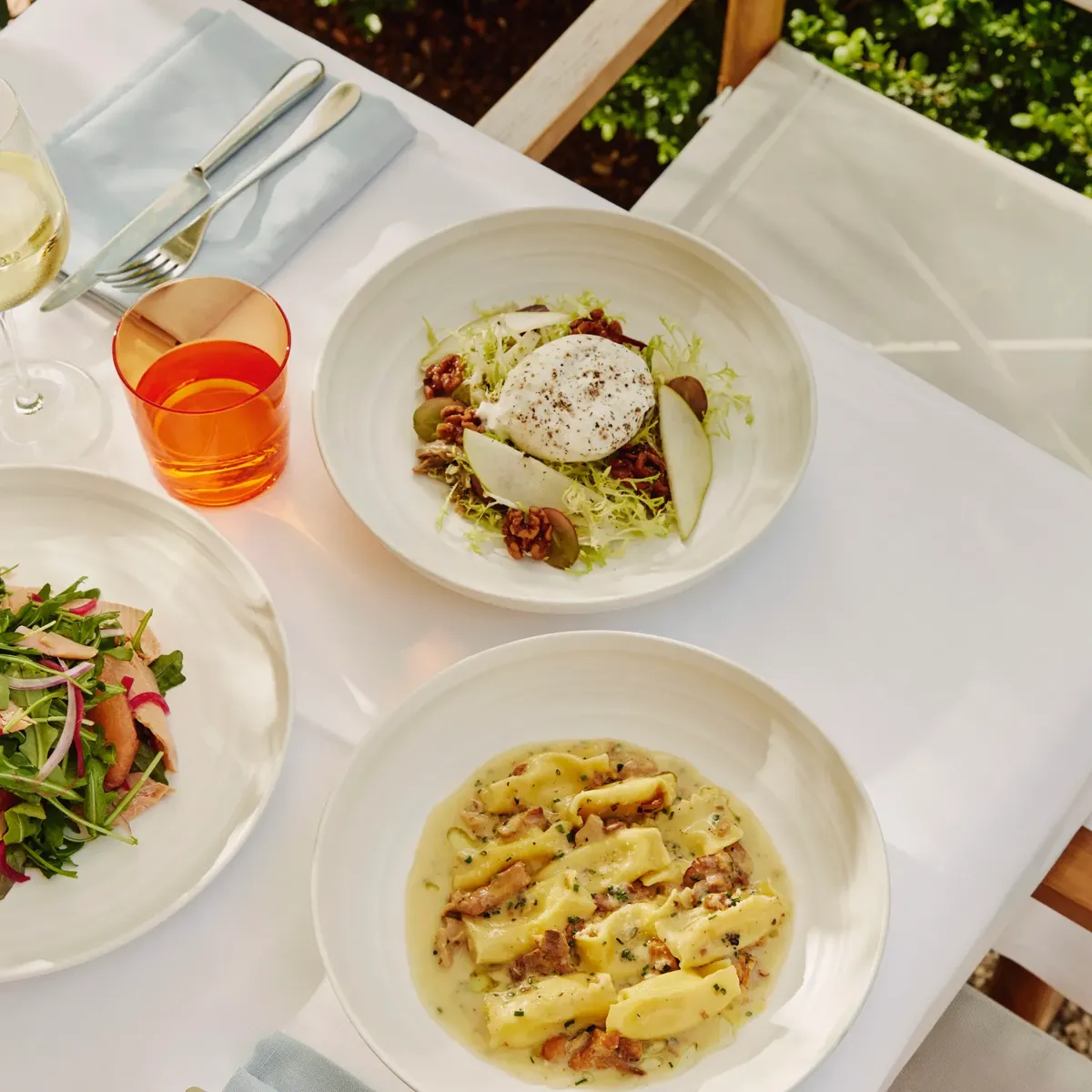 Overhead of burrata and pear salad beside gnocchi in creamy sauce on a sunlit terrace table.