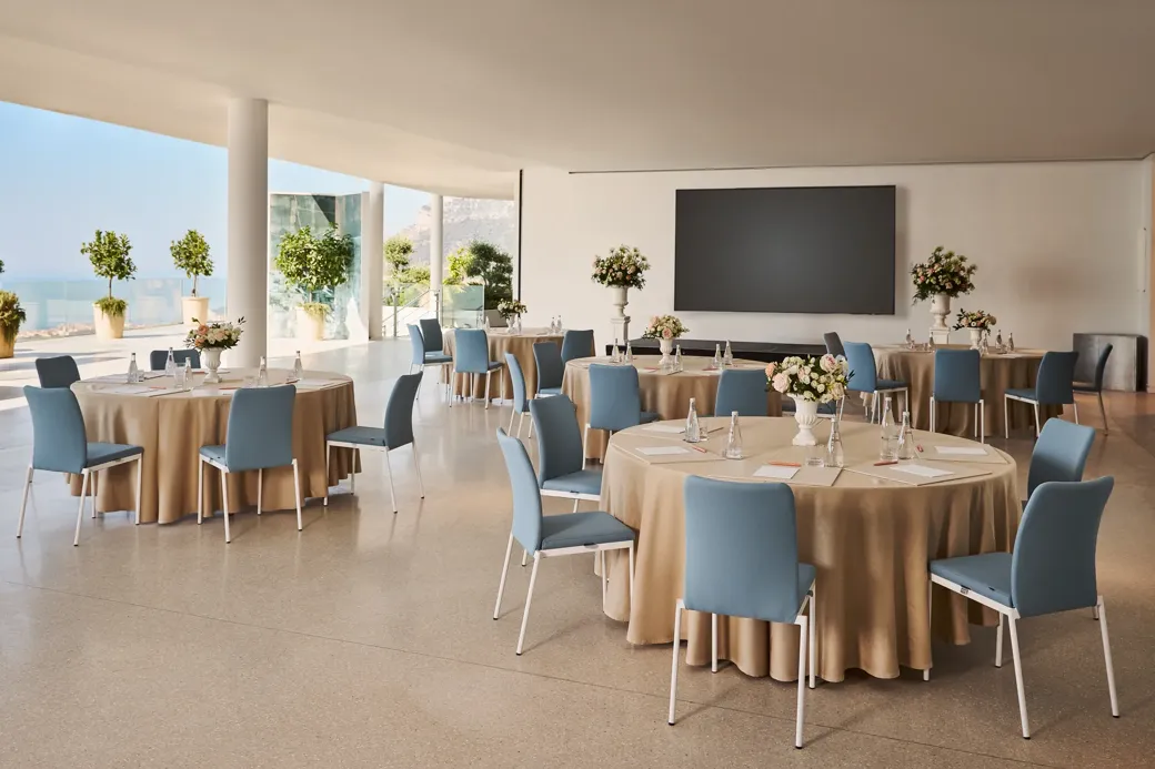 Espace événementiel intérieur avec des tables rondes recouvertes de nappes beiges, des chaises bleues, des centres de table floraux et un grand écran de présentation, offrant une vue sur la mer à travers les parois vitrées.