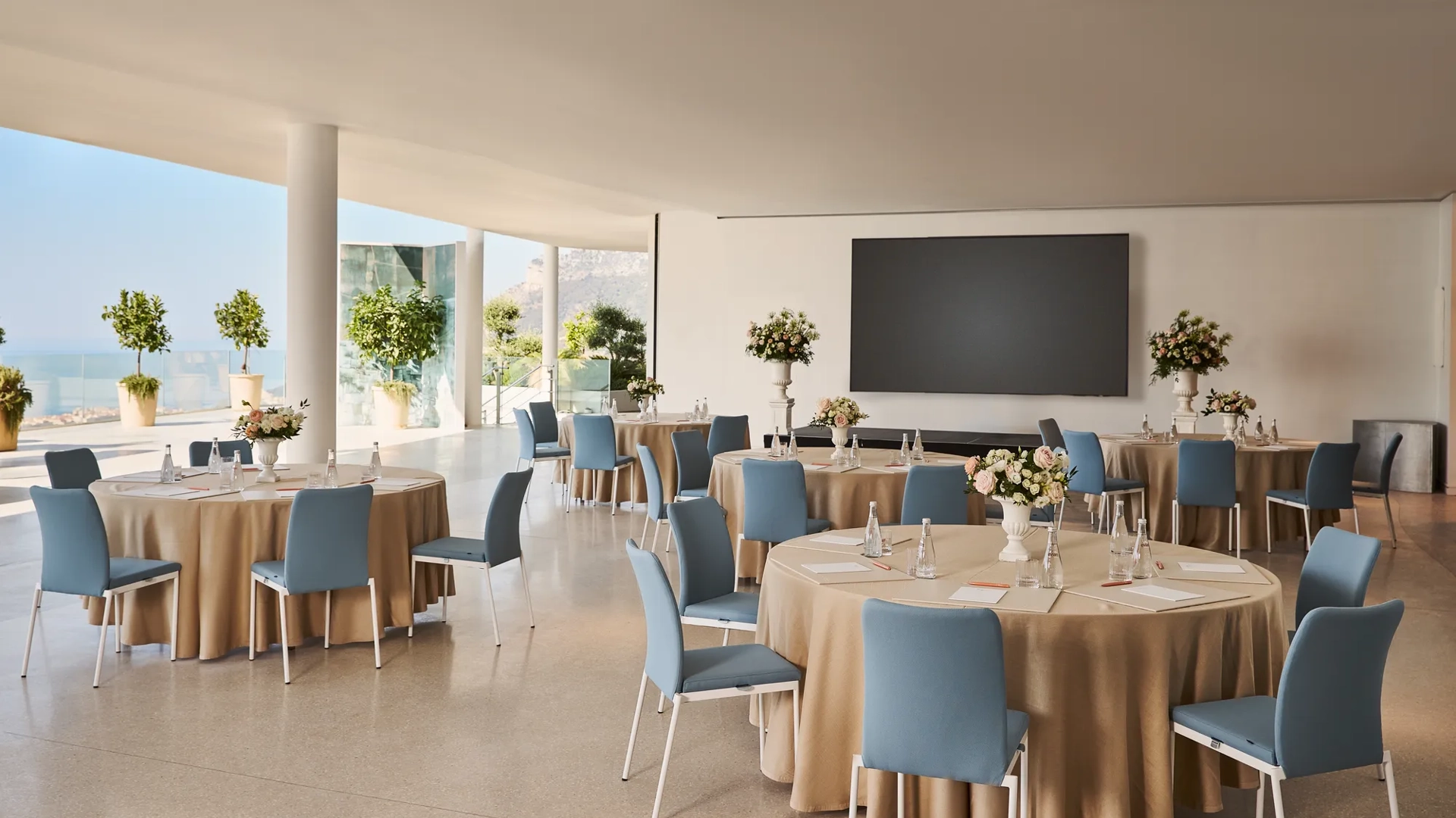 Espace événementiel intérieur avec des tables rondes recouvertes de nappes beiges, des chaises bleues, des centres de table floraux et un grand écran de présentation, offrant une vue sur la mer à travers les parois vitrées.