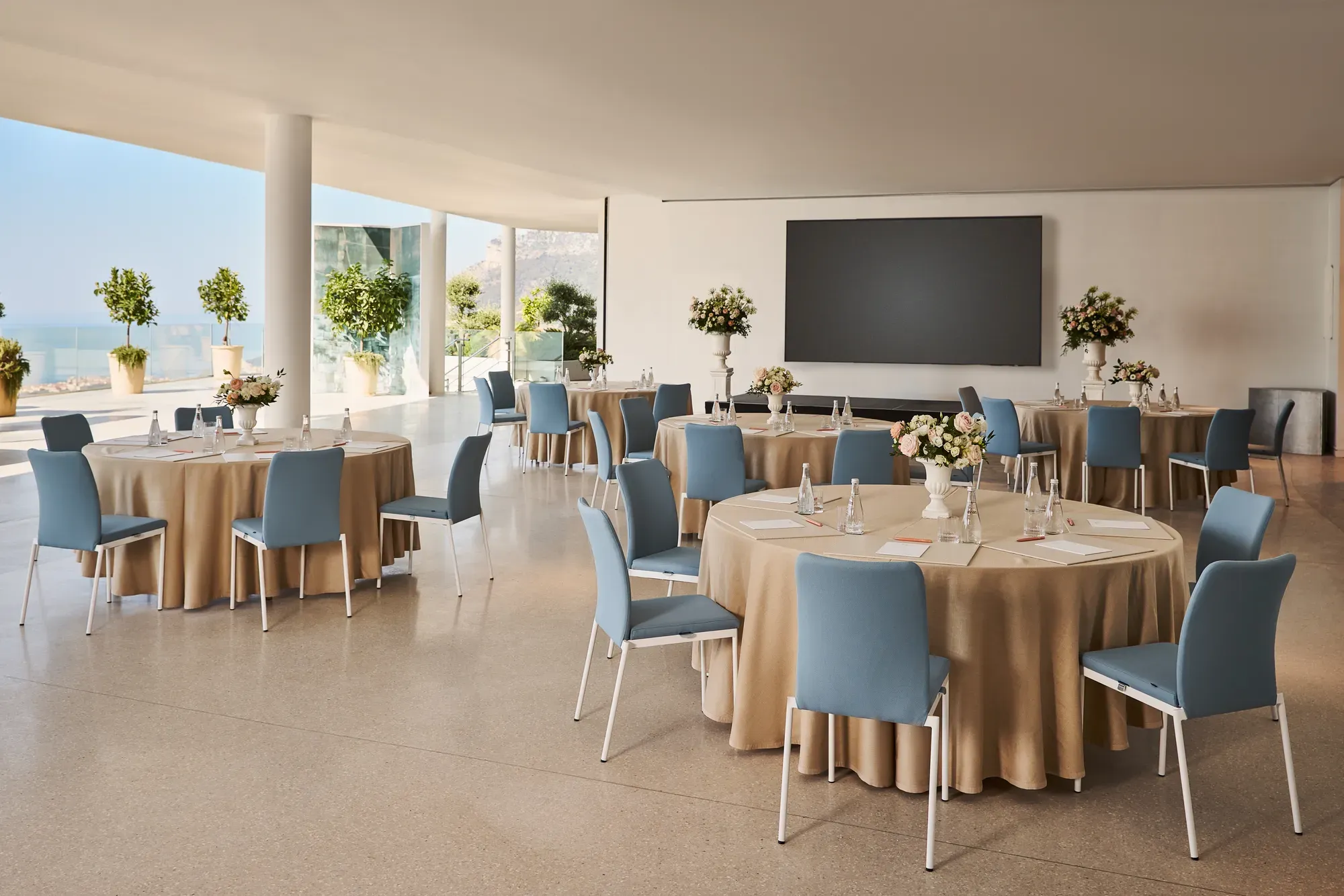 Espace événementiel intérieur avec des tables rondes recouvertes de nappes beiges, des chaises bleues, des centres de table floraux et un grand écran de présentation, offrant une vue sur la mer à travers les parois vitrées.