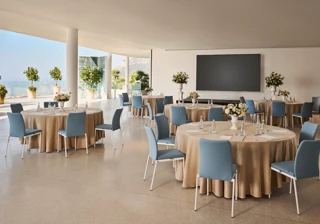 Espace événementiel intérieur avec des tables rondes recouvertes de nappes beiges, des chaises bleues, des centres de table floraux et un grand écran de présentation, offrant une vue sur la mer à travers les parois vitrées.