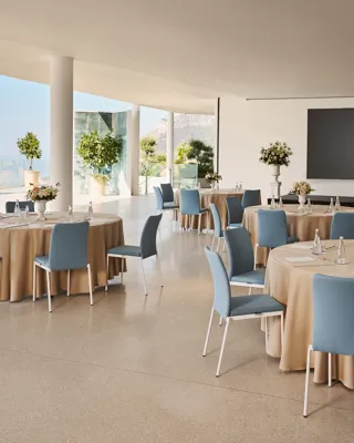 Espace événementiel intérieur avec des tables rondes recouvertes de nappes beiges, des chaises bleues, des centres de table floraux et un grand écran de présentation, offrant une vue sur la mer à travers les parois vitrées.