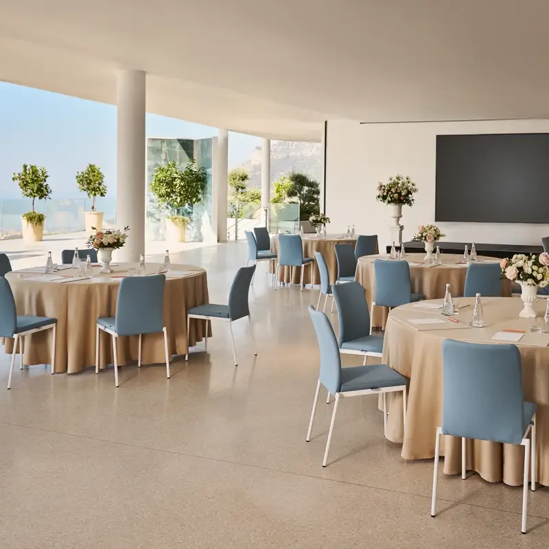 Espace événementiel intérieur avec des tables rondes recouvertes de nappes beiges, des chaises bleues, des centres de table floraux et un grand écran de présentation, offrant une vue sur la mer à travers les parois vitrées.