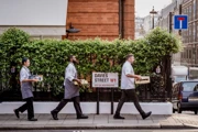 Three Claridge’s staff walk along Davies Street carrying boxes beside a green hedge.