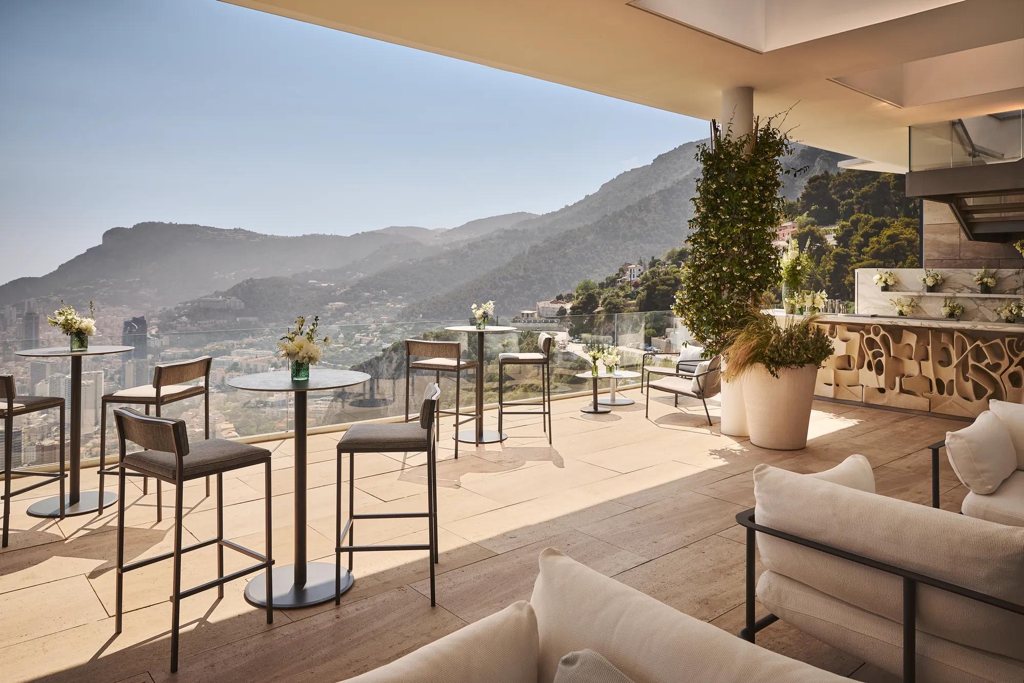 Terrasse élégante sur le toit avec tables de cocktail, bar, fleurs et vue panoramique sur montagnes et côte.