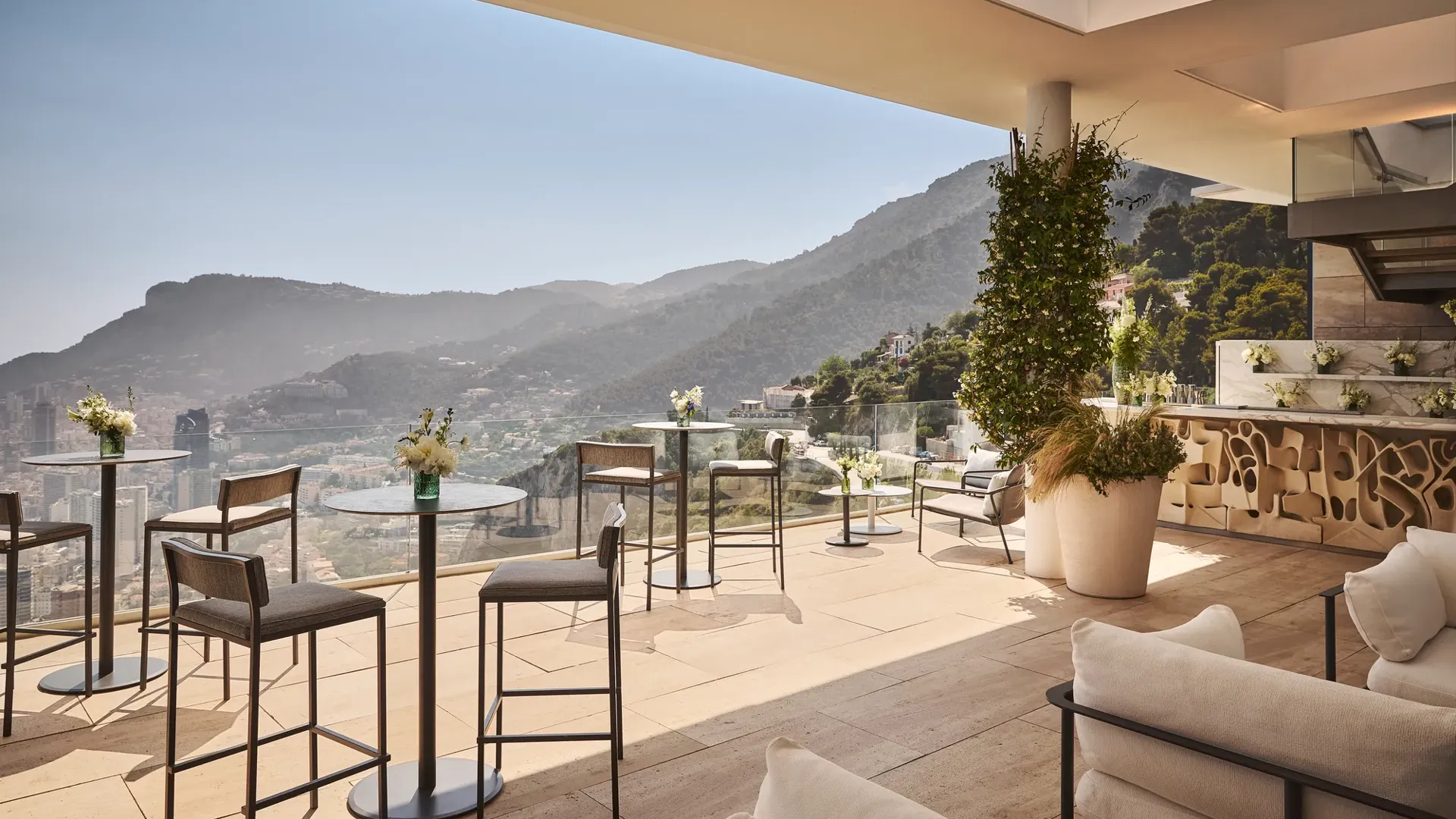 Terrasse élégante sur le toit avec tables de cocktail, bar, fleurs et vue panoramique sur montagnes et côte.