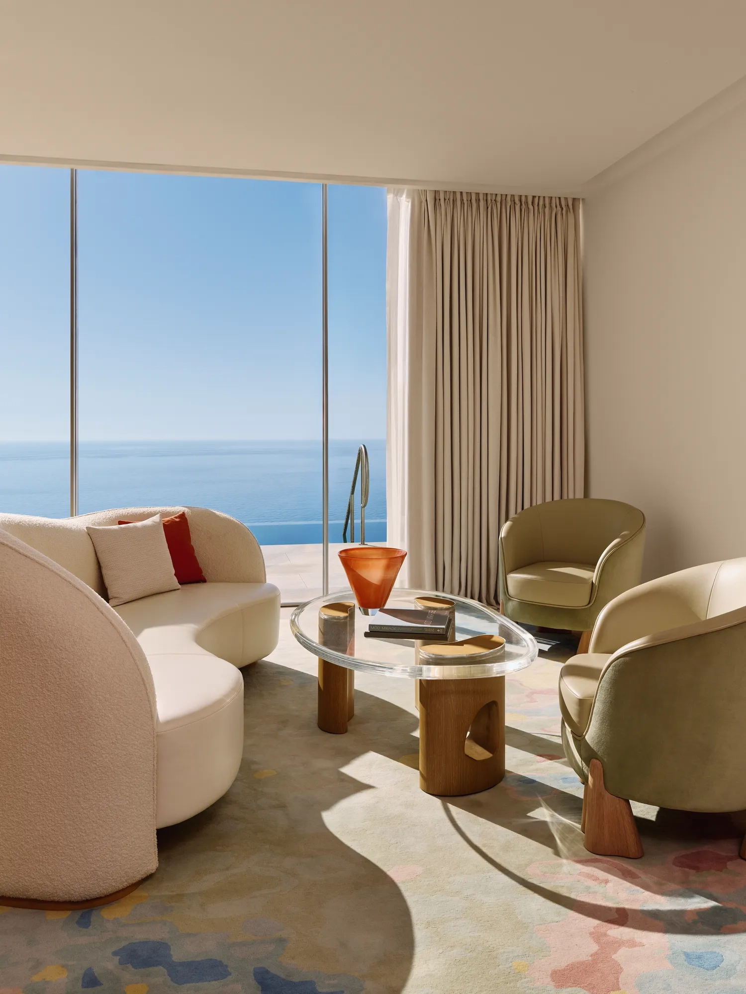 Coin salon lumineux avec canapé incurvé, deux fauteuils et table basse en verre, devant de grandes baies vitrées surplombant la mer.