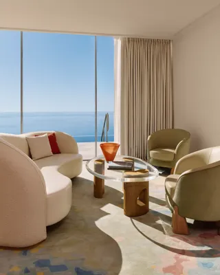 Coin salon lumineux avec canapé incurvé, deux fauteuils et table basse en verre, devant de grandes baies vitrées surplombant la mer.