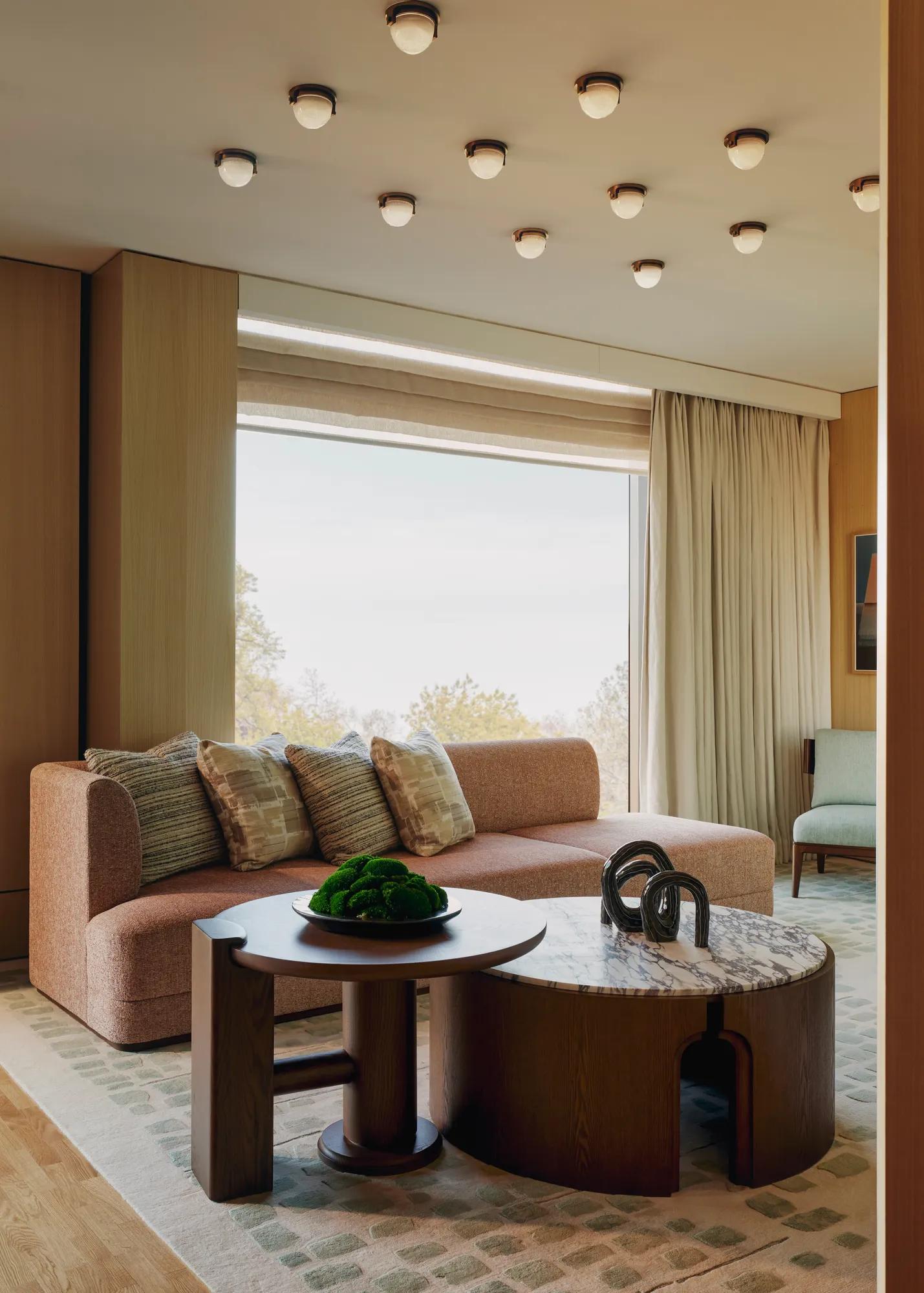 Modern suite with peach sofa, sculptural tables, and large windows overlooking trees, featuring soft lighting and warm wood tones.