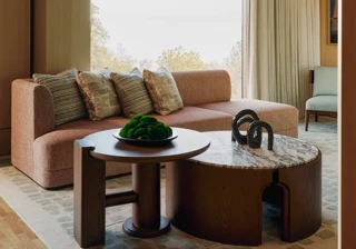 Modern suite with peach sofa, sculptural tables, and large windows overlooking trees, featuring soft lighting and warm wood tones.