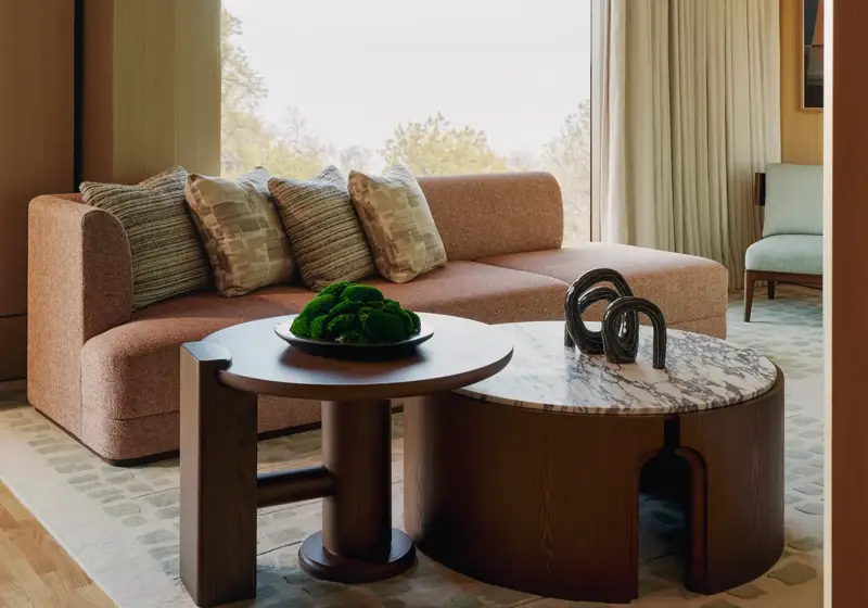 Modern suite with peach sofa, sculptural tables, and large windows overlooking trees, featuring soft lighting and warm wood tones.