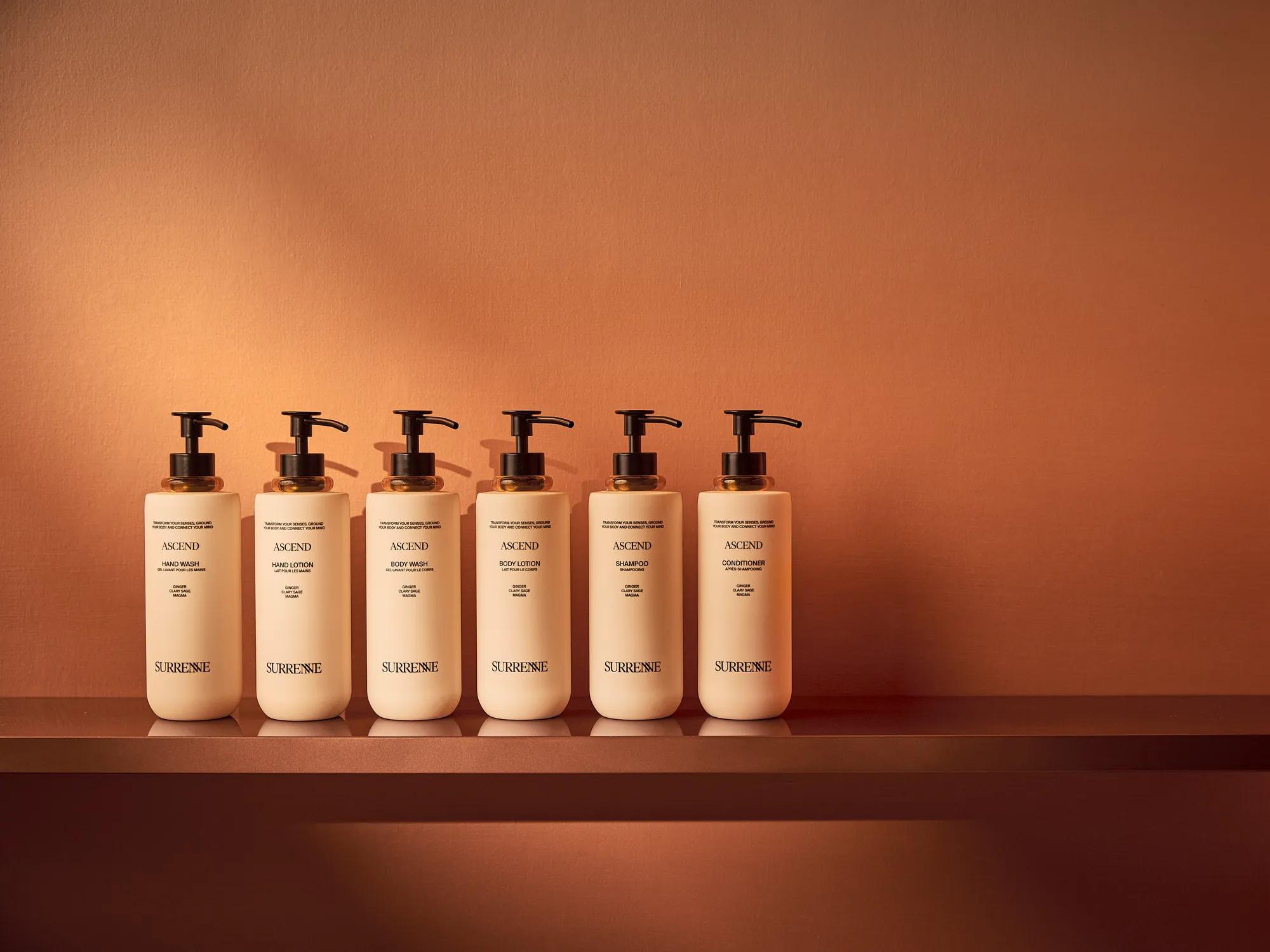 Six Surrenne Ascend bath and body bottles displayed on a shelf against a warm terracotta background.