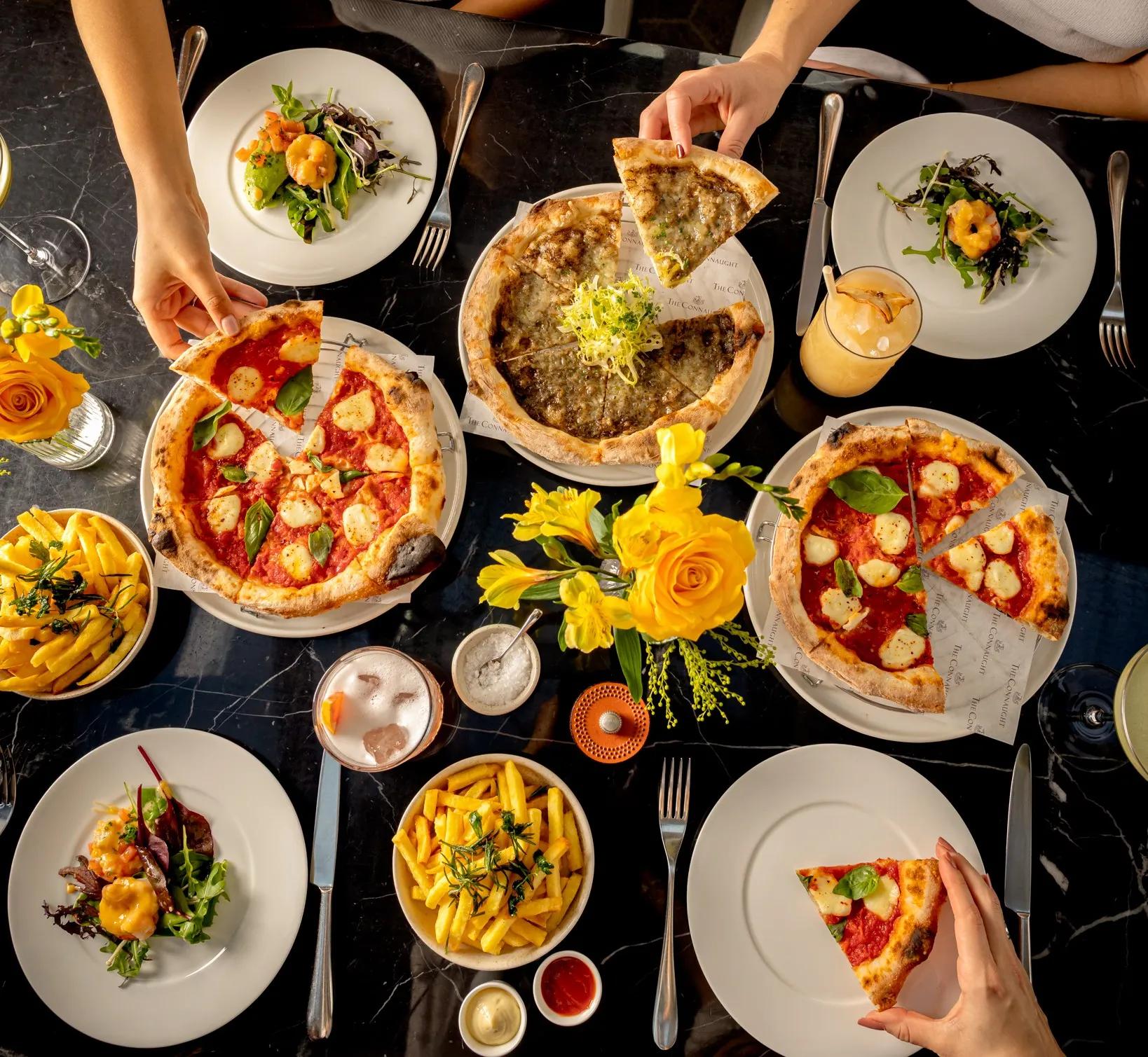 Overhead view of diners sharing pizzas, fries and cocktails on a marble restaurant table.