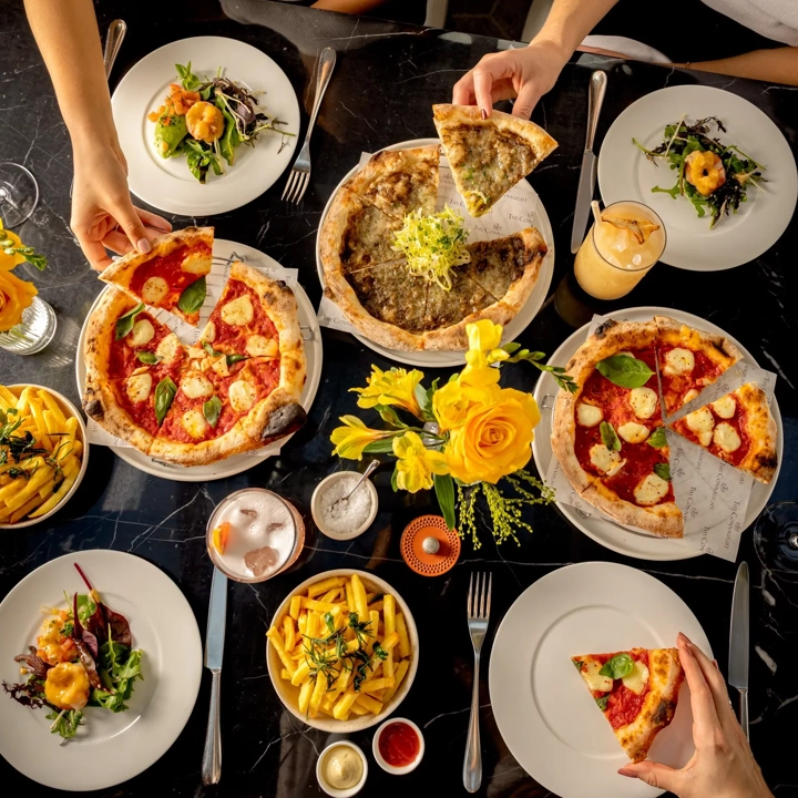 Overhead view of diners sharing pizzas, fries and cocktails on a marble restaurant table.