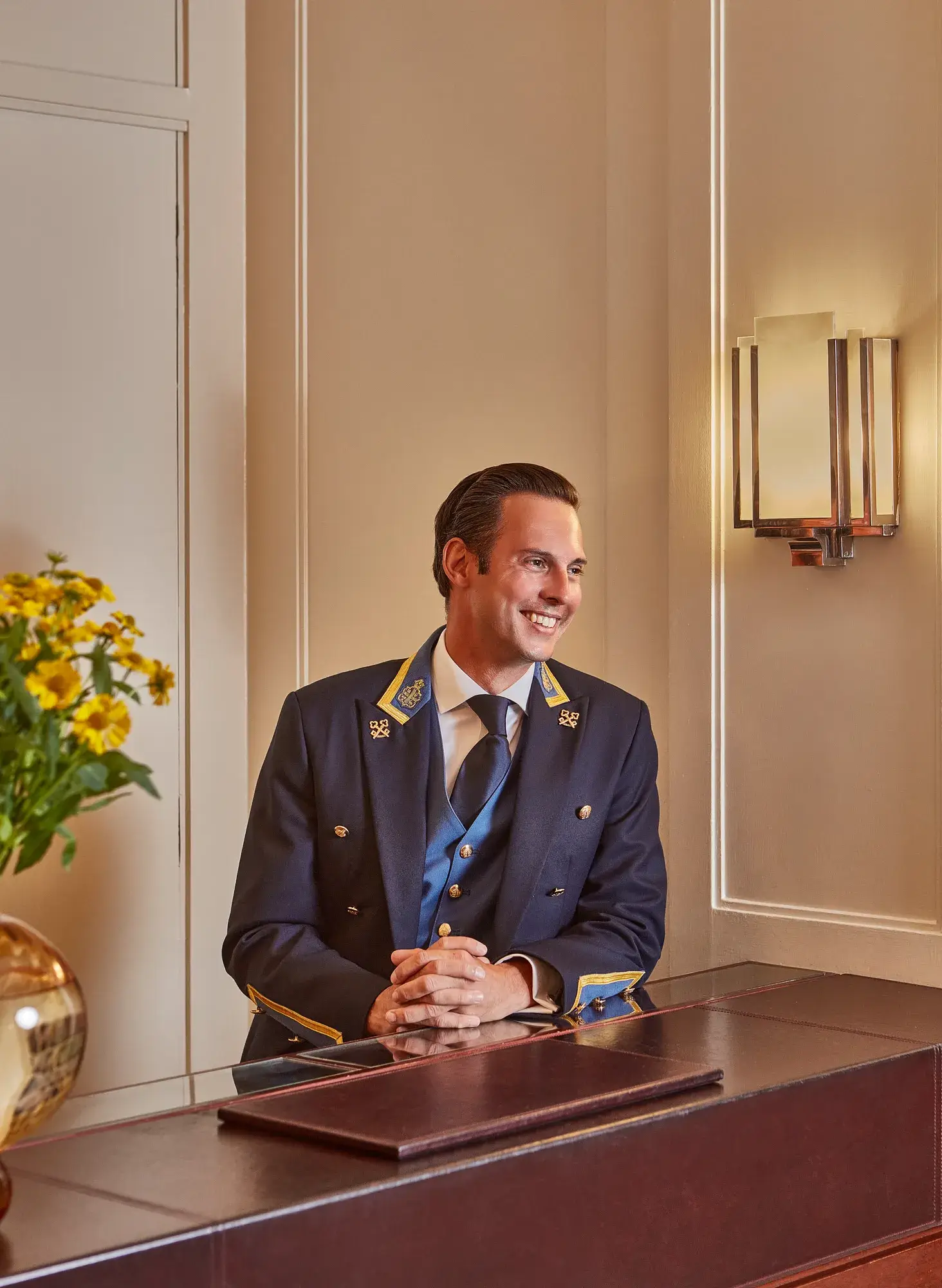 Hotel concierge in navy uniform smiling behind reception desk, with flowers and wall sconce in a warm, elegant interior.