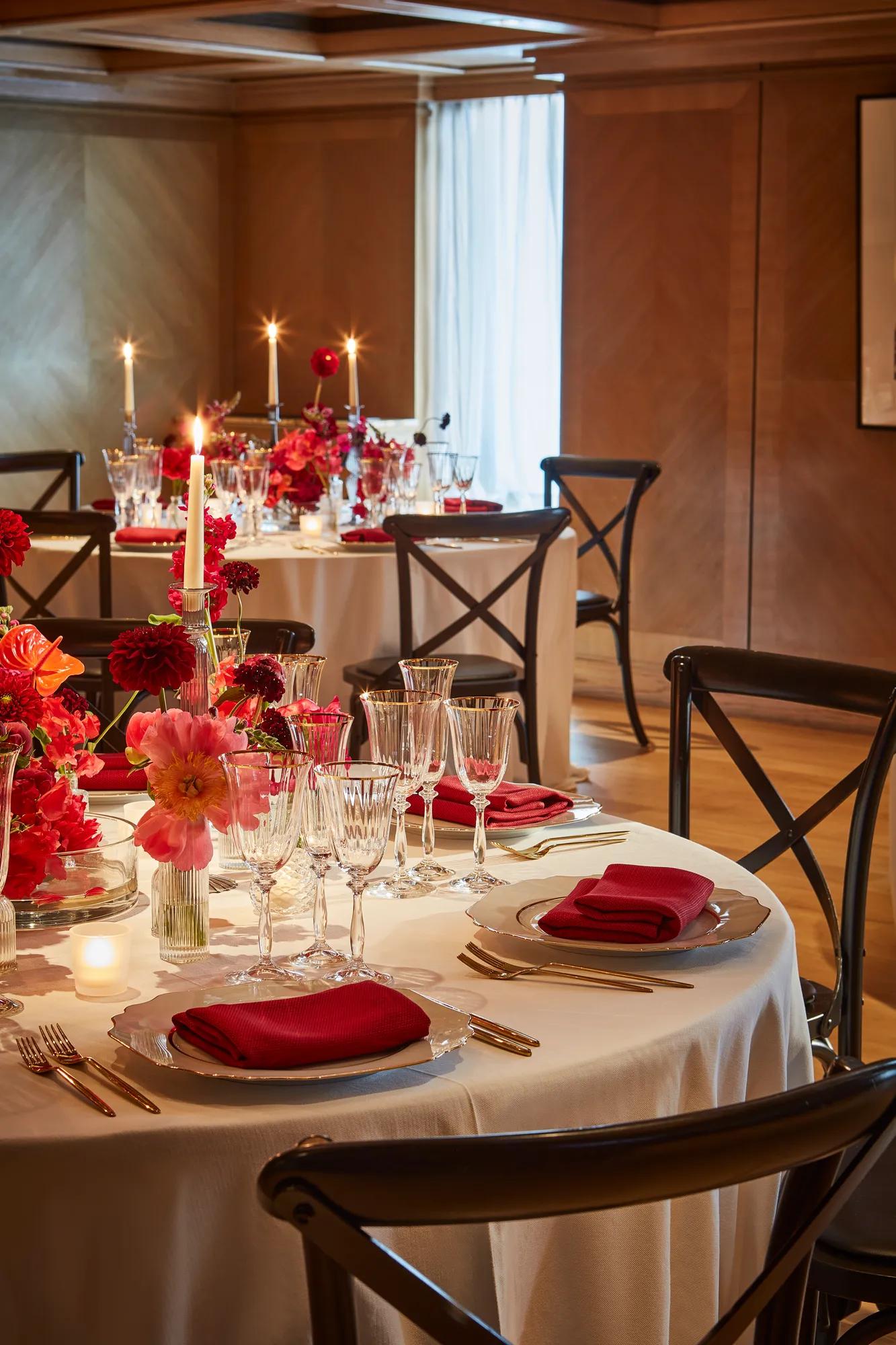 Round dining table dressed in white with red napkins, crystal glassware and vivid pink and red floral centrepieces, set with glowing candles in a warm wood-panelled room.