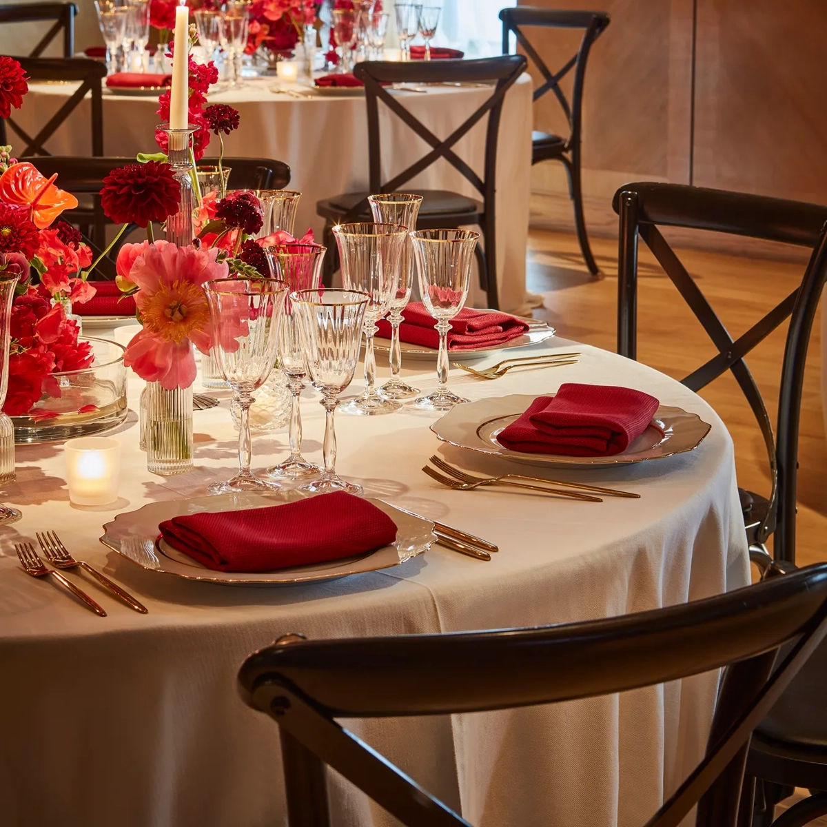 Round dining table dressed in white with red napkins, crystal glassware and vivid pink and red floral centrepieces, set with glowing candles in a warm wood-panelled room.