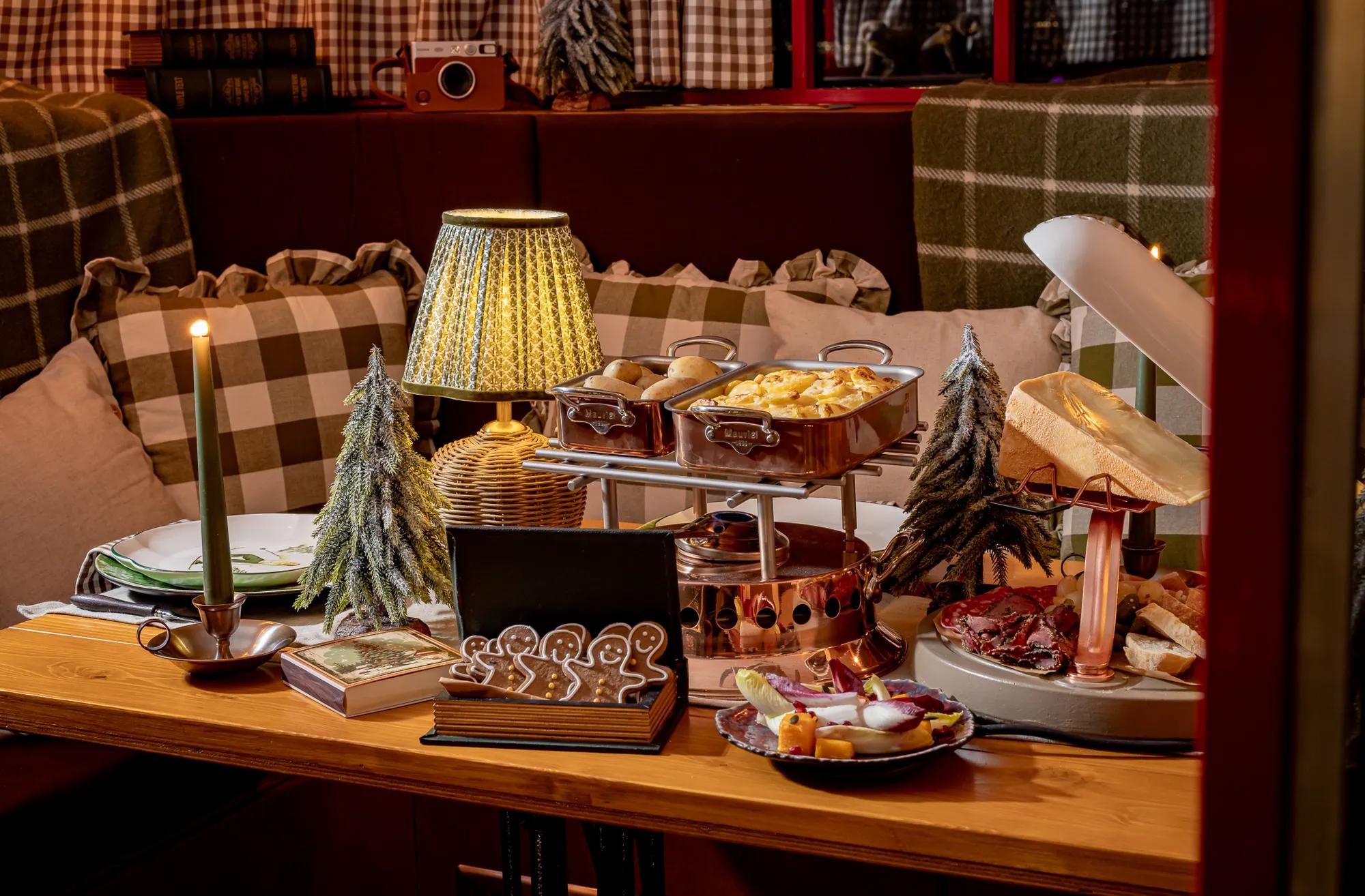 Festive dining table with raclette, charcuterie, gingerbread biscuits and miniature snowy trees, surrounded by checked cushions and warm candlelight.