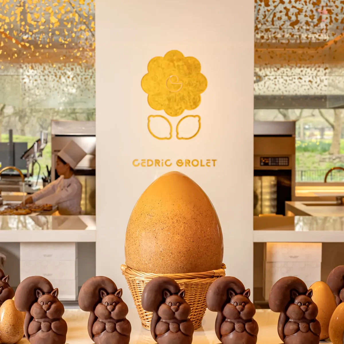 Chocolate squirrel figurines before a giant speckled Easter egg at Cedric Grolet pâtisserie counter, chef working behind.