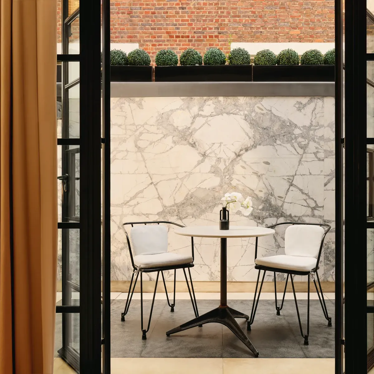 Light-filled sitting area with two upholstered chairs, round table with flowers, patterned carpet, and balcony view through tall windows.