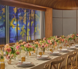 Long dining table set for an event, decorated with pink and peach flowers and glowing amber glassware