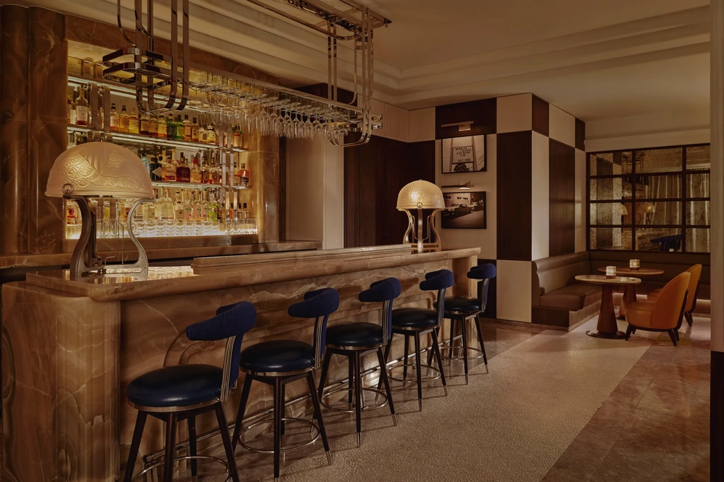 Wide view of The Maybourne Bar with onyx counter, row of navy stools, and booth seating in warm, low light.