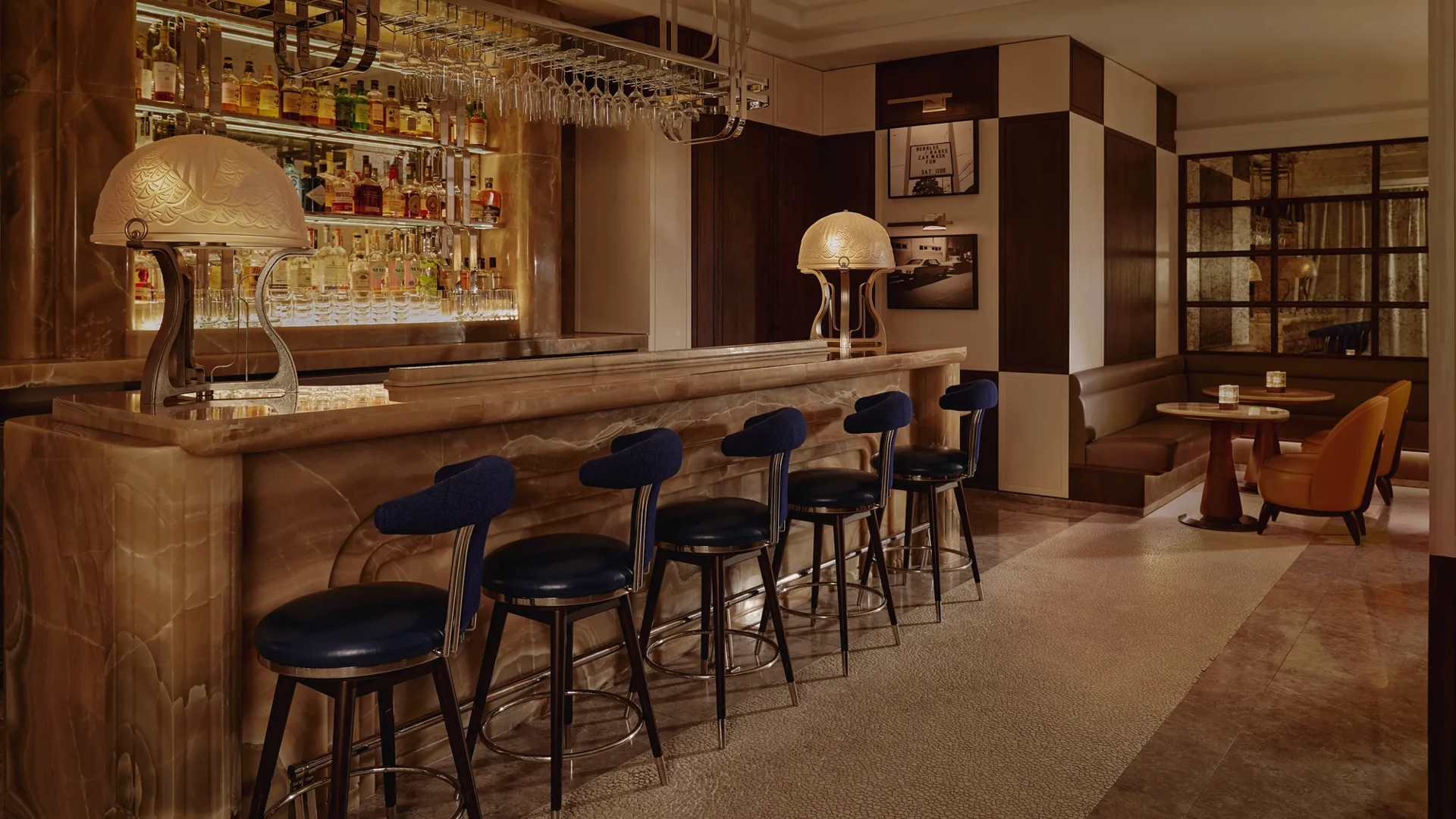 Wide view of The Maybourne Bar with onyx counter, row of navy stools, and booth seating in warm, low light.