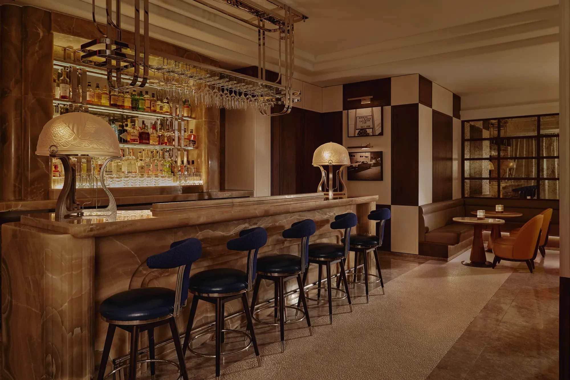 Wide view of The Maybourne Bar with onyx counter, row of navy stools, and booth seating in warm, low light.