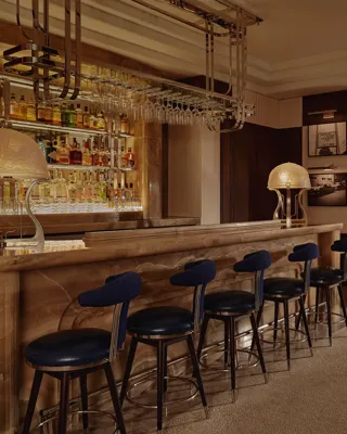 Wide view of The Maybourne Bar with onyx counter, row of navy stools, and booth seating in warm, low light.