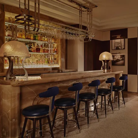 Wide view of The Maybourne Bar with onyx counter, row of navy stools, and booth seating in warm, low light.