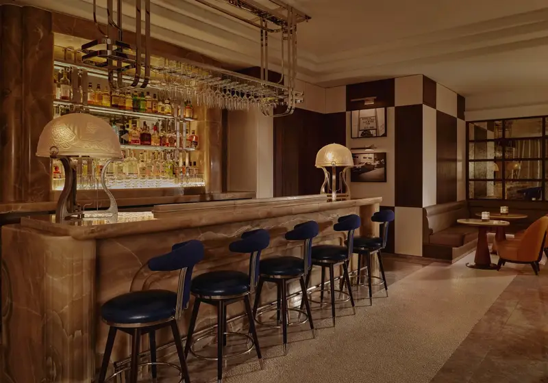 Wide view of The Maybourne Bar with onyx counter, row of navy stools, and booth seating in warm, low light.