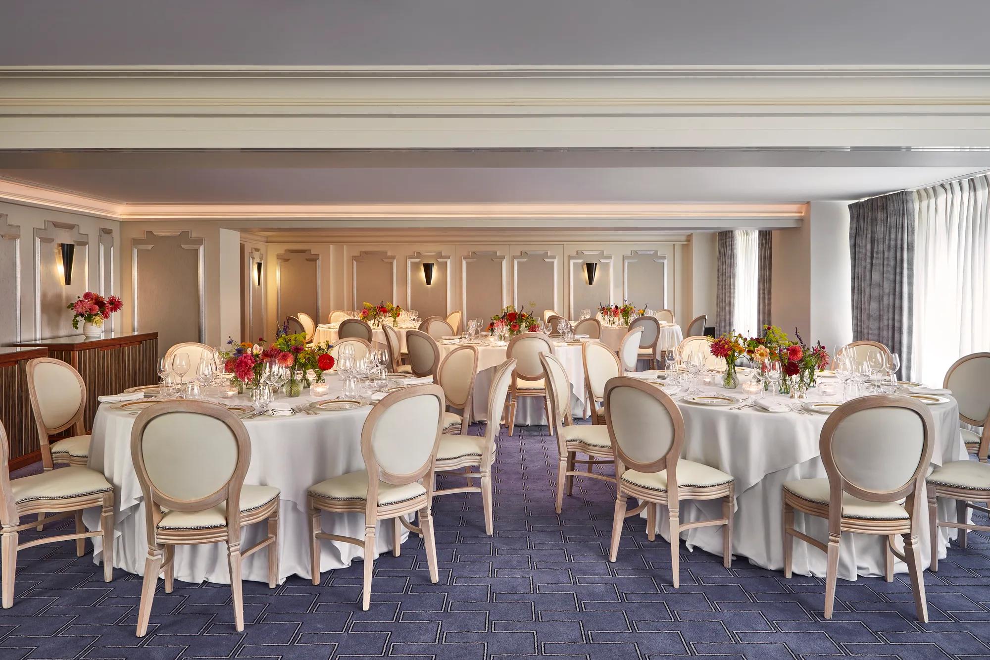 Elegant banquet room at Claridge’s set for an event with round tables dressed in white linen, cream chairs, and vibrant floral centrepieces, lit by natural light through curtained windows.