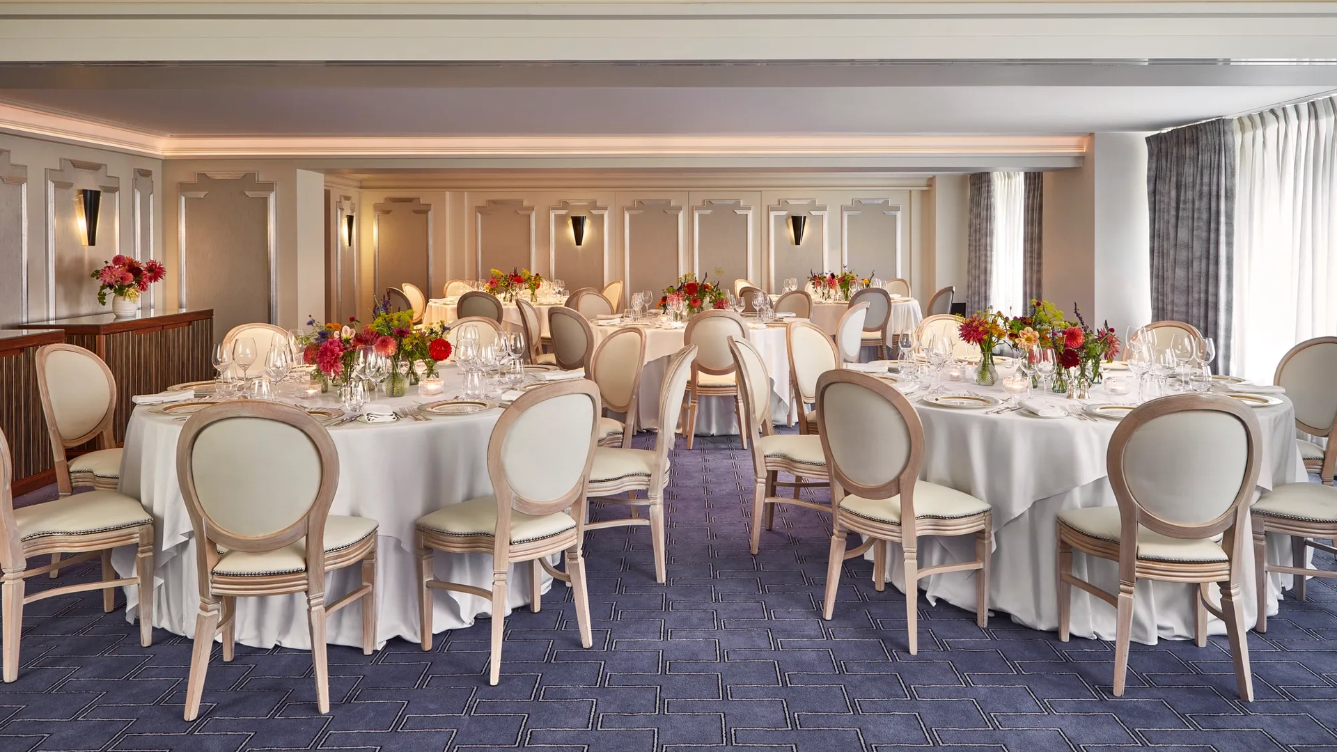 Elegant banquet room at Claridge’s set for an event with round tables dressed in white linen, cream chairs, and vibrant floral centrepieces, lit by natural light through curtained windows.