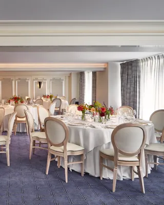 Elegant banquet room at Claridge’s set for an event with round tables dressed in white linen, cream chairs, and vibrant floral centrepieces, lit by natural light through curtained windows.