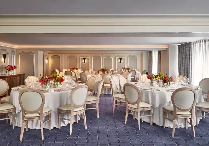 Elegant banquet room at Claridge’s set for an event with round tables dressed in white linen, cream chairs, and vibrant floral centrepieces, lit by natural light through curtained windows.