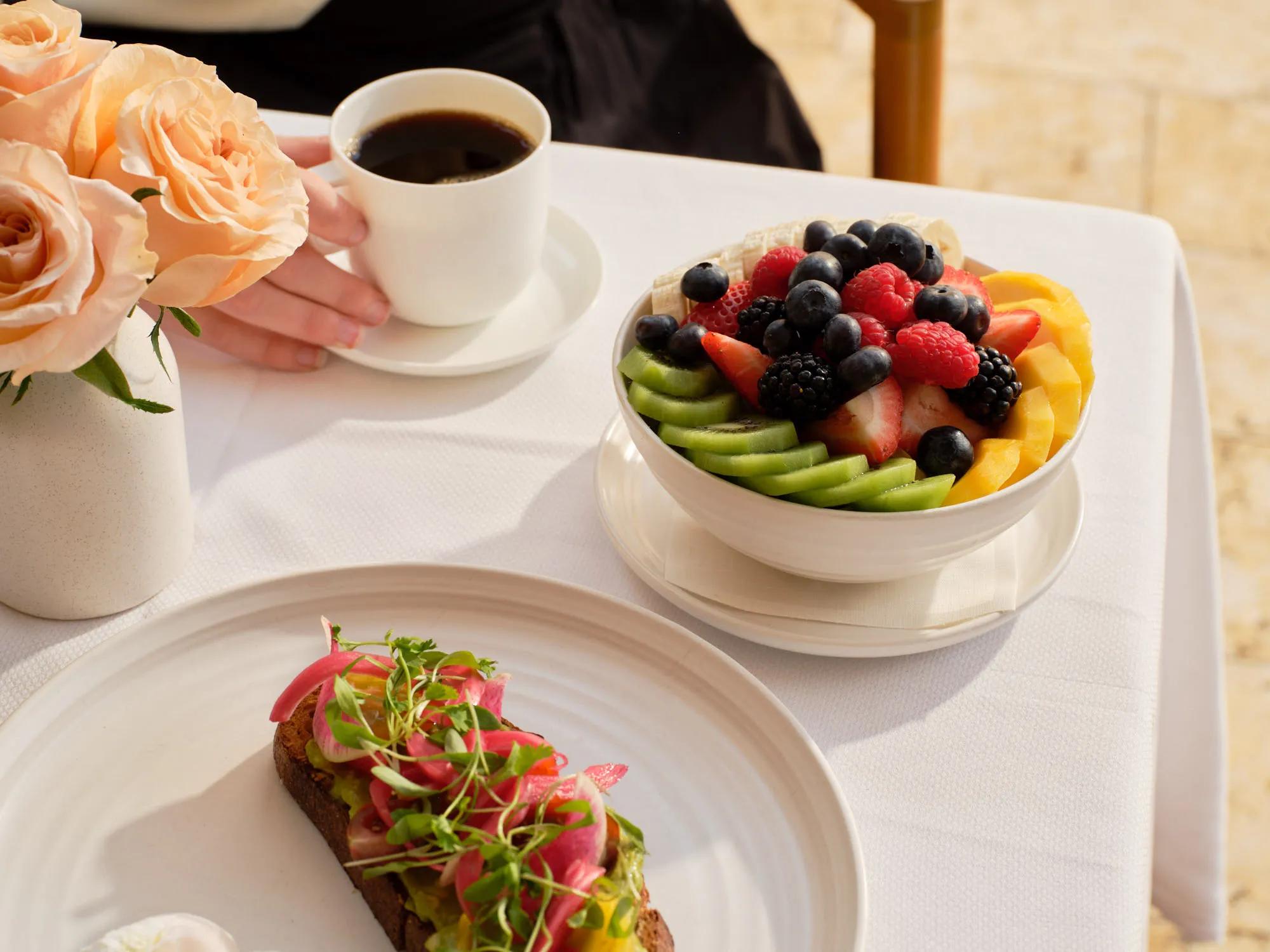 Bowl of fresh fruit with berries, kiwi, and mango, served beside coffee and avocado toast on a sunlit table with flowers.