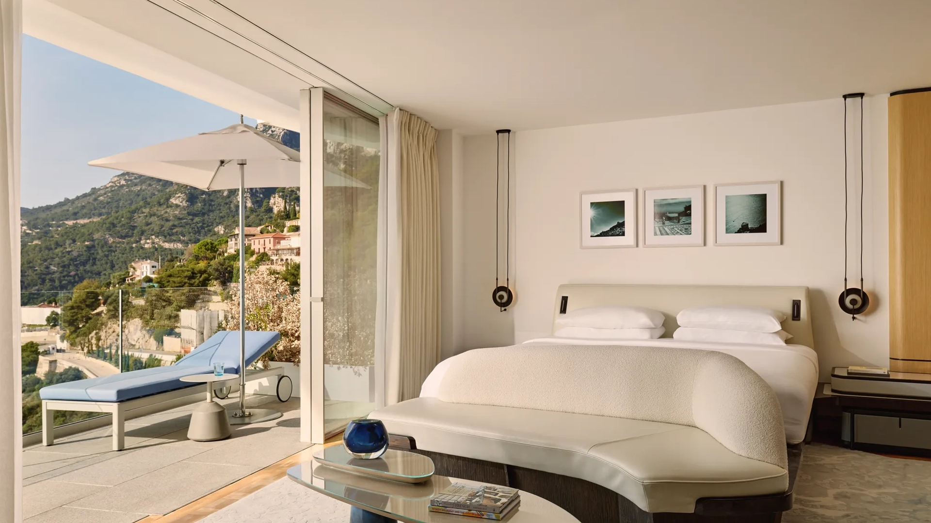 Chambre d’hôtel élégante avec grand lit, banquette incurvée et portes vitrées s’ouvrant sur une terrasse avec transat, parasol et vue sur la montagne.